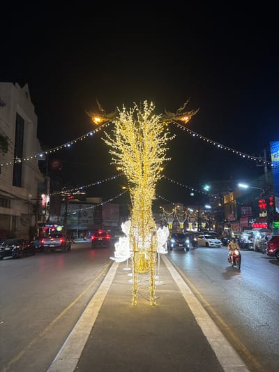 The image displays a nighttime street scene within Chiang Rai City Municipality, Thailand. A large, illuminated, deer-shaped sculpture constructed from warm white LED string lights, resembling a stylized tree, stands centrally in the road's median. Several white, illuminated lotus-like structures are positioned at the base of the sculpture.
The asphalt road features traffic lanes on both sides of the median. On the left, multiple dark-colored cars and pickup trucks are visible, moving away from the viewer. On the right, several vehicles are present, including a dark SUV, a white sedan, a red car, and a single individual operating a red scooter, all traveling towards the viewer.
Buildings line both sides of the street, featuring multi-story structures with various commercial establishments. Visible signage includes a red "Pizza Hut" logo on a building to the left and a partial blue sign, likely for a bank, on the right. The street is extensively lit by string lights draped across the thoroughfare and by ornate, traditional Thai-style street lamps. The sky is dark, indicating a nocturnal setting.