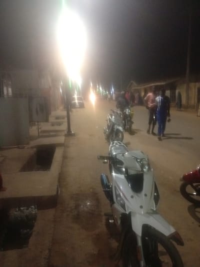 The image depicts a street scene at night in Tundun Wada West, Nigeria, illuminated by a bright streetlight. In the foreground, a white motorcycle is parked. Behind it, several other motorcycles are visible, some with riders. A group of people are standing and walking along the street. Buildings line the road, and a drainage ditch is present along the sidewalk. The overall impression is that of a busy street at night.