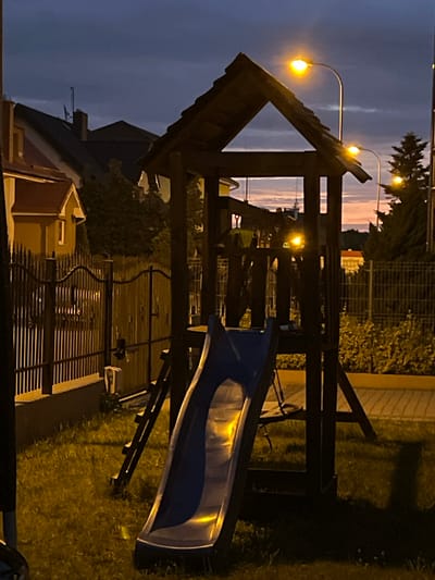 This is a vertical, medium shot of a wooden playground structure with a blue slide at dusk in Rumia, Poland. The sky is a deep blue with streaks of orange and pink from the setting sun. Streetlights cast a warm yellow glow on the scene.
The playground structure is in the foreground, dominating the center of the image. It is made of dark wood and has a peaked roof. A ladder leads up to a platform. The blue slide is attached to the platform and curves downwards onto the grass.
To the left of the playground is a yellow house with a red roof, partially obscured by trees. A black metal fence with decorative elements runs along the property line. To the right of the playground, more trees and bushes are visible, as well as another fence. The ground is covered in green grass and some pavement. The overall impression is one of a quiet, residential area at the end of the day.