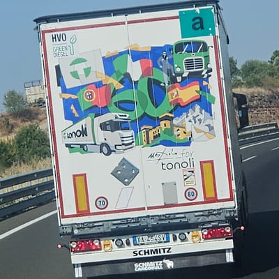 The image shows the rear of a white truck trailer with a colorful mural painted on it. The mural depicts several elements including flags of various countries (Portugal, Italy, Spain), mountains, buildings, a green truck, and a silhouette of a person. There is text on the trailer that reads "HVO GREEN DIESEL" and "tonoli". There are also speed limit signs (70 and 80). The truck is on a highway. In the foreground, the license plate of the truck is visible, which reads "XA-434SV". Below the license plate, "SCHMITZ" and "SCHMITZ CARGOBULL" are visible. The environment appears to be a rural landscape with dry vegetation and a clear sky. The specific location of Cervera, Spain is not evident from the image itself, as it is a generic highway scene.