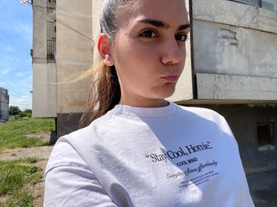 A young woman with dark hair pulled back in a ponytail and dark eyebrows is captured in a selfie outdoors. She is wearing a white t-shirt with black text that reads: "Stay Cool, Homie." Below that, "COOL MIND" and "Everything flows effortlessly." At the bottom of the text, it says: "WHEN YOUR MIND IS AS COOL AND SHARP AS ICE AND THE ATMOSPHERE IS RIGHT, NOTHING CAN THROW YOU OFF." She is standing in front of a Brutalist-style apartment building with a weathered facade and visible signs of disrepair. The building is pale beige and gray, with some sections appearing more worn and damaged than others. The sky above is blue with scattered white clouds, and there are some faint contrails visible. To the left of the building, there is a patch of green grass and weeds, with a dirt path. The overall setting appears to be a residential area in a city. The photograph was taken in the city of Debelt, Bulgaria.