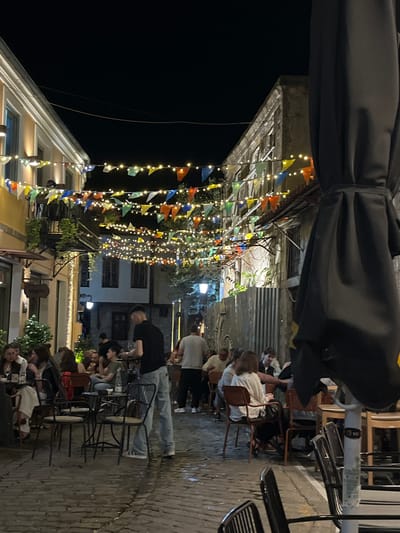 This image captures a street scene in Xanthi, Greece, at night. Overhead, strings of colorful pennant flags and fairy lights are strung between buildings. People are seated at outdoor cafe tables, socializing and eating. The street is paved with stone. Several people are wearing casual clothing. In the foreground, a partially visible black umbrella canopy dominates the top part of the frame. Tables and chairs are spaced along the street.
