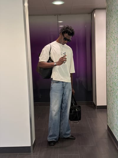 A full-body, medium-shot, indoor, selfie shows a Black male, appearing to be in his early twenties, standing in a hallway with a purple, gradient-style door and a tiled wall. The subject is wearing a loose-fitting, off-white t-shirt, light-wash, wide-leg jeans, and black, pointed-toe loafers. He has dreadlocks, is wearing black sunglasses, and is holding a silver smartphone in his right hand, taking the picture. A black, leather-style shoulder bag is slung over his left shoulder, and a second black duffel bag is held in his right hand, resting on his hip. The hallway floor is tiled with dark gray square tiles, and the walls are a mix of white and a green, mosaic-style tile. The lighting suggests overhead fixtures, casting a soft glow. The location is identified as Crawley, United Kingdom.