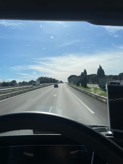 The image displays a daytime scene from the driver's perspective inside a vehicle traveling on a multi-lane highway near Tavel, France. The lower portion of a dark gray steering wheel and a dark dashboard occupy the foreground, with a dark rectangular device mounted to the lower right. The windshield shows sunlight glare and minor smudges. The asphalt highway, marked with white lines, recedes into the distance under a bright blue sky with scattered white clouds. Several vehicles are visible, including a dark passenger car in the left lane ahead and a white semi-truck further back. Metal guardrails separate the highway from the verges. The surrounding environment features green vegetation, including dense treelines on the left and distinctive tall, conical trees mixed with rounded trees on the right. No human figures are explicitly visible.