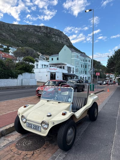 A light-colored dune buggy is parked on the brick-paved sidewalk, close to a manhole cover. Behind the buggy is a red car parked on the street, in front of a building with the sign "Pizza Shed" on it. The building behind the Pizza Shed is a light blue, multi-story structure. A street lamp stands tall in front of the building. In the background is a large mountain, and a partly cloudy blue sky is visible above it. In the upper part of the background, near the building, some people are seen standing on the sidewalk. Based on the architecture, location, and signage, this is likely a street scene in Muizenberg, South Africa.