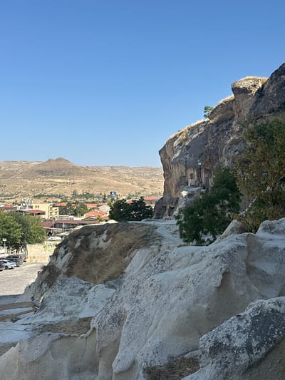 The image shows a landscape in Ürgüp, Turkey, characterized by its unique geological formations and a town nestled within the hills. In the foreground, a close-up view reveals the textured, pale grey rock formations, typical of the region's tuff stone. These rocks appear weathered and sculpted by erosion, with some areas showing dry grasses and sparse vegetation.
Moving beyond the immediate foreground, the scene opens up to a broader panorama. A prominent cliff face, also made of tuff, dominates the right side of the image. Within this cliff, several carved openings and niches are visible, suggesting historical human habitation or religious sites. A single green tree grows on a ledge of this cliff, adding a splash of color against the arid landscape. Further back, a path or road winds its way up the rocky terrain.
In the middle ground, the town of Ürgüp is visible. Buildings with red-tiled roofs are clustered together, interspersed with trees. Some of the structures appear to be modern, while others might be older, carved into the rock. A prominent yellow building stands out among the others. Beyond the town, the landscape transitions into rolling, arid hills, dotted with sparse vegetation and possibly some man-made structures or power lines in the distance. The sky above is a clear, bright blue, indicating a sunny day.
In the lower left portion of the image, a paved area resembling a parking lot is visible, with several cars parked. A low stone wall and a wooden railing suggest a viewing area or a pathway along the edge of the precipice. The overall impression is one of a vast, ancient landscape shaped by both nature and human history.