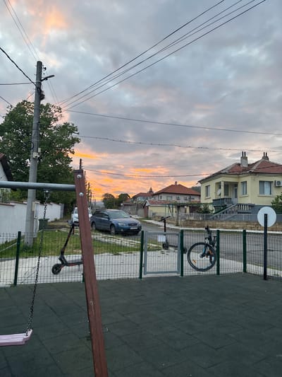 The image depicts a street scene in Goritsa, Bulgaria, at sunset. A row of residential buildings lines the street, including a two-story yellow building with a satellite dish. A grey car is parked along the side of the street. Power lines stretch across the sky. There is a playground area with a fence, rubber matting, a bicycle parked against the fence, and a scooter nearby. Part of a swing set and a brown bench is in the foreground.
