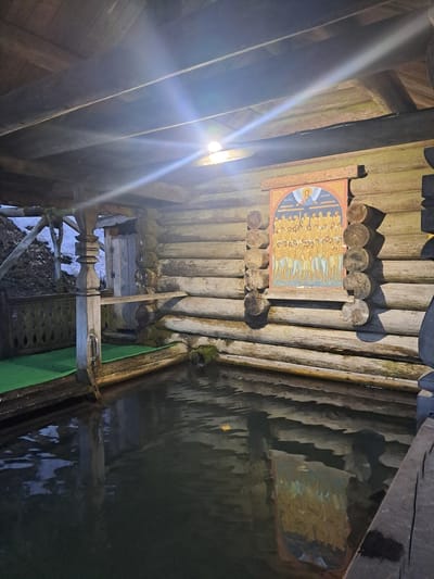 The image depicts the interior of a wooden structure, likely a wellspring or small building, located near Vzglyadnevo, Russia. A religious icon is affixed to the log wall, held in place by wooden posts. Water is present in the structure, reflecting the surrounding wooden walls and roof. A ladder or steps leads down into the water. Debris, including leaves and snow, are visible near the base of the steps. A green covering is positioned near the water's edge. Artificial light illuminates the scene from the left side.
