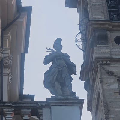 The image shows a low-angle view of a stone statue of a robed figure with a feathered headdress, positioned between two ornate architectural structures. The statue is centrally located and appears to be standing on a pedestal. To the left of the statue is a cream-colored architectural element with curved details and vertical fluting. To the right, a stone tower or belfry is visible, featuring rows of bells and decorative stonework. The sky in the background is overcast and bright. This statue is located in the Piazza IV Novembre in Calcinate, Italy, atop the Loggia Civica.