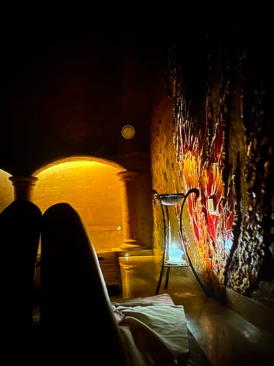 The image shows a dimly lit interior space, possibly a spa or relaxation room. In the foreground, two dark silhouettes of legs are visible, draped with a light-colored towel or sheet. The legs are positioned as if resting.
The environment features an arched entryway with illuminated stone columns on either side, casting a warm yellow light. Beyond the archway, a textured wall with what appears to be a mosaic or decorative panel is illuminated by a light source, creating a vibrant red and orange glow. A slender, dark metal stand with a shallow bowl sits in front of this illuminated wall.
The overall ambiance is tranquil and serene, with the low lighting and warm colors suggesting a relaxing atmosphere. The location is not specified to be in Denver, Colorado, United States, and no forensic details are discernible from the image.