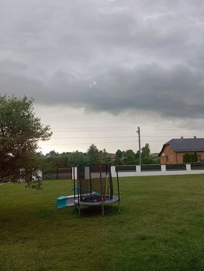 The image captures a backyard scene in Hlynytsi, Ukraine, under a heavy, overcast sky. In the foreground, a trampoline with a safety net is visible, accompanied by a blue and white slide. The lawn is green and well-maintained, with some scattered small white flowers. A mature tree with dense foliage stands to the left. In the mid-ground, a low white fence with horizontal dark elements separates the yard from a slightly elevated area with more trees and vegetation. Beyond this, a brick house with a dark roof and red chimney pots is situated on the right, with power lines and a utility pole visible in the distance. The overall impression is of a rural or suburban residential area on a cloudy day.