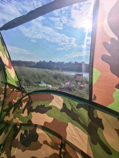 The image is a view from inside a camouflage tent looking outwards. Through the mesh window of the tent, a body of water, possibly a river or lake, is visible in the distance, with a tree line on the opposite bank. The sky is blue with scattered clouds, and the sun is shining brightly, creating a lens flare effect. In the foreground outside the tent, on a grassy bank, are several colorful items, including a pink object, a blue object, and a green bag. A folding camping chair is also visible. The tent material itself, with its camouflage pattern of green, brown, and black, occupies the foreground of the image. The overall scene suggests an outdoor recreational setting, likely a campsite near a body of water. The location is identified as the village of Kuzminovka, Russia.