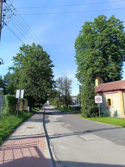 The image depicts a residential street in Czekanów, Poland, on a clear, sunny day. A paved road with visible patches and repairs runs through the center of the frame, with a sidewalk on the left side. The sidewalk is constructed of light-colored pavers near the grass and red pavers closer to the street. A wire fence runs along the edge of the sidewalk, bordering a grassy area.
On the right side of the road, a small building with a tiled roof and a yellow facade is visible. A bell tower is attached to the roof. Next to the building, a speed limit sign indicating "30" within a red circle is posted. Tall, mature trees with dense green foliage flank both sides of the street, casting shadows on the road and sidewalk. Power lines crisscross the sky, supported by a concrete pole on the left. The sky is bright blue with a few wispy clouds. In the distance, houses and more trees can be seen lining the street. No people or vehicles are present in the immediate foreground.