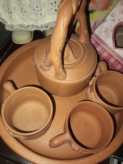 A terracotta tea set is displayed on a matching tray. The set includes a teapot with a sculpted handle resembling intertwined branches and three cups, each with a small handle. Floral patterns are carved near the handles of the cups. The teapot sits centrally on the tray, surrounded by the cups. A porcelain doll and a patterned box are partially visible in the background.