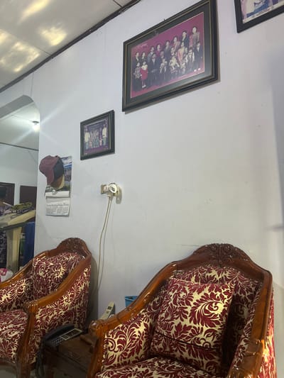 The image depicts an indoor scene, likely a living room in Terang-terang, Indonesia. Two ornate wooden chairs with red and gold patterned upholstery are prominent in the foreground. A small wooden table between the chairs holds a corded telephone and what appears to be a cordless phone base. On the white wall behind the chairs, two framed photographs are displayed. The larger, horizontally oriented photograph at the top features a group of people in formal attire, including children. A smaller, vertically oriented photograph is positioned below it. To the left of the photographs, a calendar with a scenic image hangs on the wall, adorned with two caps, one maroon and one brown. A power outlet with a plug is also visible on the wall. The ceiling has a textured finish with light patterns cast upon it, suggesting light sources from above or through a window. The overall impression is of a well-furnished, residential interior.