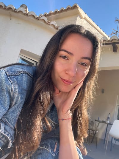 A young woman with long brown hair poses for a selfie. She is wearing a blue denim jacket and has a red bracelet on her wrist. Her right hand is resting on her face. The background shows a building with white walls and a tiled roof under a clear blue sky. In the distance, a table and chairs are visible. The location is likely el Poble Nou de Benitatxell / Benitachell, Spain, based on the architectural style.