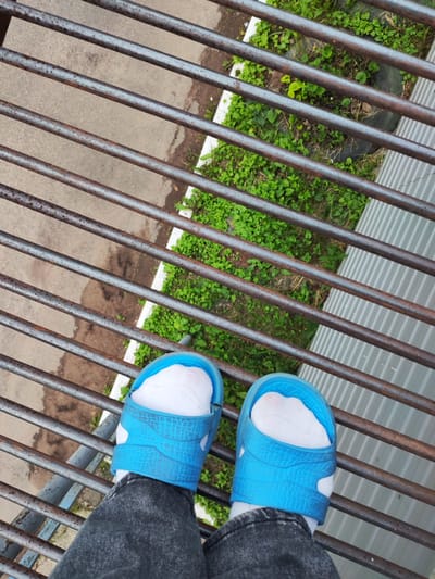 The image shows a top-down view of a person's feet wearing blue sliders and white socks, standing on a rusted metal grate. The grate is positioned over a patch of green vegetation and a grey surface, possibly pavement. The environment appears to be outdoors, with hints of an urban or semi-urban setting due to the metal structure and the ground surface. The lighting suggests it is daytime. The location in the city of Borodino, Russia, is not visually discernible from the image itself and would require external context to confirm.
