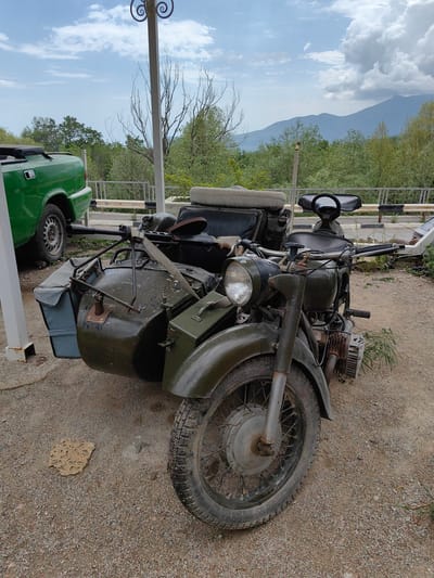 The image presents a vintage military motorcycle with a sidecar, located in what appears to be an outdoor display or parking area. The motorcycle is painted in a dark olive green color and exhibits signs of wear and age. The tire in the front is worn, indicating some use. The sidecar is attached to the left of the motorcycle, and it also shows its age.
The surrounding environment includes a gravelly ground surface, some vegetation, and parts of a green vehicle. The sky is partly cloudy, and mountains are visible in the background. Based on the prompt, the location is Luchistoe, Ukraine.