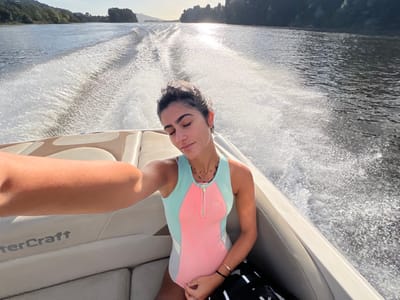 The image depicts a female subject in a boat on a body of water in Vila Nova de Cerveira, Portugal. The subject, an adult female with dark hair styled away from her face, is positioned on the starboard side of a moving powerboat, partially facing the camera while extending her right arm. Her eyes are closed, and her head is slightly tilted. She is wearing a sleeveless, color-blocked swimsuit in mint green, peach, and white/light gray, featuring a visible zipper and a logo on the chest. Accessories include a dark choker necklace, a delicate gold necklace, a gold bracelet on her right wrist, and a black band on her left wrist. A black and white striped object, possibly a bag or towel, rests beside her left hip.
The boat's interior features light-colored, upholstered seating with "WaterCraft" visible on the side paneling. The boat is actively moving, generating a distinct white wake behind it that extends into the background. The body of water, likely the Minho River, appears dark with shimmering reflections of sunlight in the distance. Lush, green, tree-covered landmasses form the distant banks under a clear sky. The scene is illuminated by bright natural sunlight, creating a glare on the water surface in the far background.