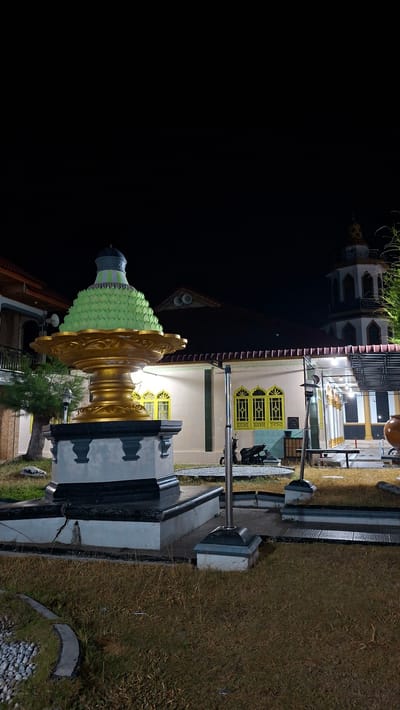 The image depicts a night scene in Lhokdagang, Indonesia, featuring a large, ornate golden fountain-like structure with a layered, green, conical top in the foreground. The fountain is situated on a multi-tiered, dark-colored base with grey accents. Behind the fountain, a mosque with a tall minaret is visible, illuminated by artificial lights. The mosque has a white facade and distinctive yellow-framed windows. The surrounding area appears to be a courtyard or garden, with dry grass, a few trees, and what looks like a paved pathway. A motorcycle is parked near the mosque, and outdoor seating or tables are also present under a covered area. The sky is dark, indicating it is nighttime.