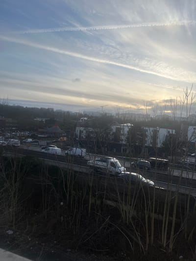 This elevated view captures an urban landscape in Rosny-sous-Bois, France, under a largely overcast sky with streaky clouds and prominent contrails, two of which intersect to form an "X." The sky brightens significantly towards the right horizon, indicative of a low sun.
In the midground, a multi-lane highway or major road runs horizontally, accommodating moderate vehicular traffic. Visible vehicles include several white vans, a silver sedan, and darker passenger cars and SUVs, moving in both directions. Adjacent to the road, on an elevated embankment, a dense thicket of bare deciduous trees and shrubs obscures the immediate foreground, with some light-colored refuse visible among the branches in the lower left.
Beyond the highway, residential or commercial buildings with dark facades and prominent white window frames or panels are visible. To the left, a distinct building features a reddish, conical roof. In the distant background, several tall construction cranes indicate ongoing development against the hazy skyline. No individuals are discernible within the frame.