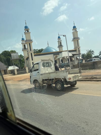 The image depicts a roadside scene in bright daylight, featuring a mosque, a small pickup truck, and a person. The mosque, a prominent structure, has a large blue dome and multiple slender minarets. These minarets are cream-colored with blue-capped tops, each adorned with a crescent moon finial. In the foreground, a white, compact pickup truck with an open bed is either parked or moving alongside the mosque. The truck's bed is fitted with a light-colored metal framework or rack. A person is visible inside the cabin of the truck, seated on the passenger side.
The setting appears to be an urban or semi-urban area, with a paved asphalt road featuring a yellow dividing line. Trees are visible behind and around the mosque, and other faint structures can be discerned in the background. The clear blue sky with scattered white clouds indicates sunny weather. Distinct shadows cast by the truck on the road suggest it is mid-day. The photo itself seems to be taken from inside another vehicle, with part of a dark window frame visible in the lower right corner. No significant text is clearly legible on the truck or other objects, though a small, blurry logo is present on the truck's door.