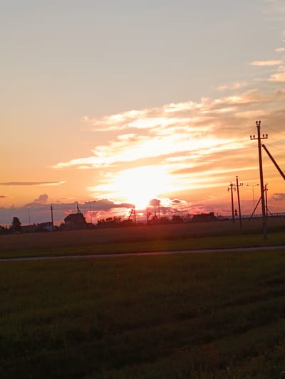 The image depicts a rural landscape at sunset, likely within the Смілавіцкі сельскі Савет, Belarus. The sky is dominated by a vibrant gradient of orange, pink, and yellow hues, characteristic of a setting sun, with scattered clouds reflecting the light. Silhouetted against this colorful sky are several utility poles with electrical lines, streetlights, and a cluster of buildings, including what appears to be a house with a steeply pitched roof. The foreground consists of a grassy field bordered by a dark road or path. The overall scene suggests a peaceful, twilight atmosphere in a semi-rural or village setting. There are no people or animals visible in the frame.