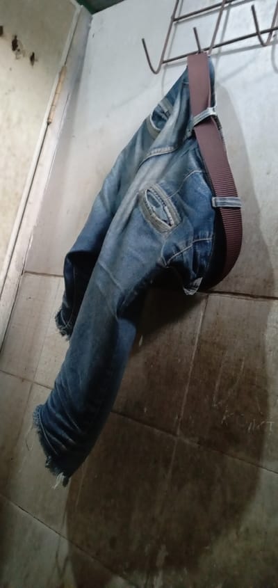 In a dimly lit room in Sumput, Indonesia, a pair of worn blue jeans hangs from a wire coat rack. A brown belt is looped through the jeans. The jeans show signs of wear and tear, with frayed edges at the bottom. The wall behind the jeans is tiled and marked with shadows, while the wall to the left appears to be painted and shows some damage. The scene is captured from a low angle, emphasizing the jeans hanging against the wall.