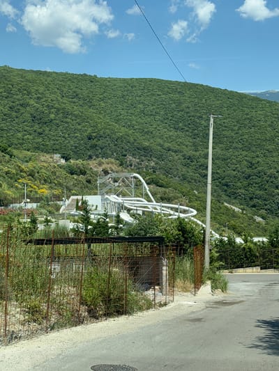 An outdoor shot captures a portion of a waterpark constructed on a lush, green mountainside under a bright blue sky with scattered white clouds. The waterpark features a complex structure of white plastic slides, support beams, and stairs, winding through the elevated terrain. The surrounding hillside is densely covered with green trees and vegetation, with some areas exhibiting yellow flowering bushes. In the foreground, a rusty metal fence encloses an overgrown area of grass and weeds, adjacent to a paved road. A tall utility pole stands to the right of the waterpark structure. The scene appears to be located in or near Podostrog, Montenegro.