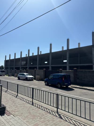 The image shows an unfinished concrete structure with exposed rebar, suggesting ongoing construction. Two cars are parked on the adjacent asphalt road, which is marked with white lines. One car is a dark blue minivan, and the other is a lighter-colored hatchback. The road is bordered by a sidewalk paved with light-colored rectangular stones. A black metal fence casts a shadow across the sidewalk and part of the road. In the background, there is a clear blue sky, indicating a sunny day. Power lines are visible in the upper left corner of the image. The scene appears to be in an urban or semi-urban environment. No people are visible in the image. There is some text visible on the license plate of the blue car, which appears to read "BKC 042 BE". The location context provided is Sarny, Ukraine.
