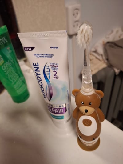 This is a close-up, horizontal shot of a bathroom sink area, with a focus on dental hygiene products. The primary subjects are a children's toothbrush with a teddy bear-shaped handle and a tube of Sensodyne toothpaste. The toothbrush has a clear plastic handle with a brown teddy bear head at the end, featuring small black eyes and a nose. The bristles of the toothbrush are visible and appear slightly used. The tube of toothpaste is white with blue and purple accents and text. Visible text on the toothpaste tube includes "HALEON," "NEW," "WORLD'S #1 SENSITIVITY TOOTHPASTE BRAND," "SENSODYNE," and "CLINICAL REPAIR." There are also mentions of "STARTS TO REPAIR SENSITIVE TEETH IN 2 MIN." A partially visible green bottle, likely another personal care product, is in the foreground. The background is slightly blurred, showing what appears to be a tiled surface or countertop and possibly fabric. The lighting suggests an indoor setting, likely during the day given the overall brightness. There is no indication of people or a specific event, but the items suggest routine personal hygiene. The location context provided is "Ogre, Latvia," but this information is not visually evident from the image itself.