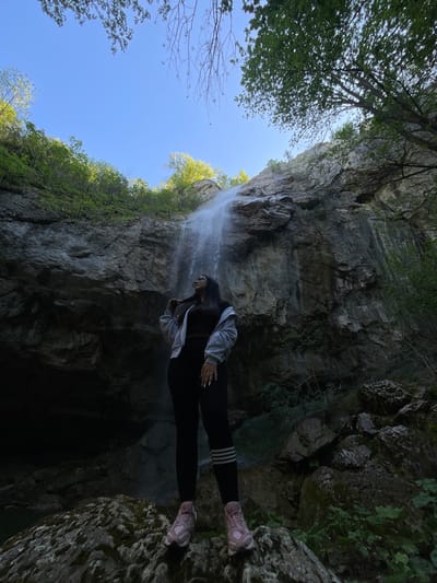 The image shows a low-angle shot of a woman standing at the edge of a cliff in what appears to be a gorge or ravine. A waterfall cascades down the rock face behind her. The sky is visible above, framed by the rocky walls of the gorge and some trees. The woman is wearing a grey jacket, black pants with white stripes, and pink sneakers. The location appears to be near Veselinovo, Bulgaria, based on the landscape and potential presence of waterfalls and rocky formations in the area.