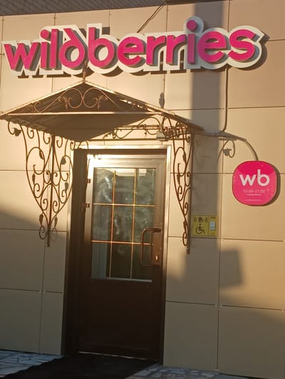 The image shows the entrance to a Wildberries store. A large pink and white "wildberries" sign is mounted above a dark brown door. To the right of the door is a circular pink sign with "wb" in white text, below which are the operating hours "10:00-21:00" and "ежедневно" (daily) in smaller white text. Below the circular sign is a yellow sign depicting a wheelchair symbol and a button, indicating an accessible entrance. The building's exterior is clad in beige paneling. The scene is illuminated by sunlight, casting shadows on the building and the ground. The location is identified as Prokudskoye, Russia.