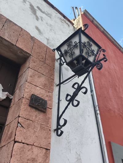 The image captures a street scene in Querétaro, Mexico. The façade of a building is visible, constructed with pink stone and white plastered walls. An ornate, black wrought-iron street lamp is affixed to the wall, featuring a decorative cage around the light source. A small wooden plaque displaying the number "129" is attached to the stone wall near a dark wooden door, partially obscured by a white cloth hanging from the door. A red-colored wall is visible on the right side of the image, with white pipes running along it. The sky is blue and clear in the upper part of the frame.
