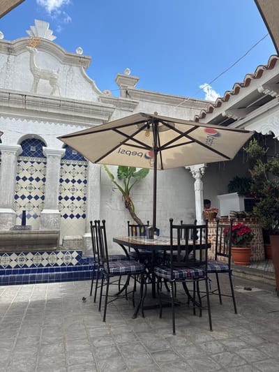 Outdoor seating area with a table, chairs, and a Pepsi umbrella. The area features ornate architecture, including a white building with decorative elements and a blue-tiled staircase. A plant grows near the table. The sky is visible, suggesting it's an open-air courtyard or patio.