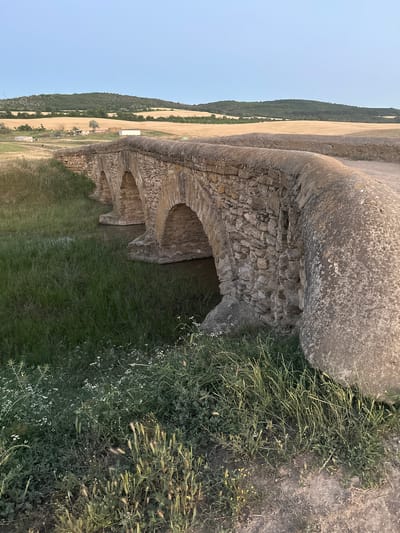 The image depicts an ancient stone bridge with multiple arches spanning a small body of water. The bridge is constructed from rough-hewn stones, showcasing signs of age and weathering. Lush green grass and vegetation grow along the banks of the water and around the base of the bridge. In the background, rolling hills are visible, covered in fields of dry, golden crops, suggesting a rural landscape. The sky above is a pale blue with a hint of dusk. The scene appears to be a serene, natural setting, emphasizing the timeless quality of the stone structure against the backdrop of the countryside.