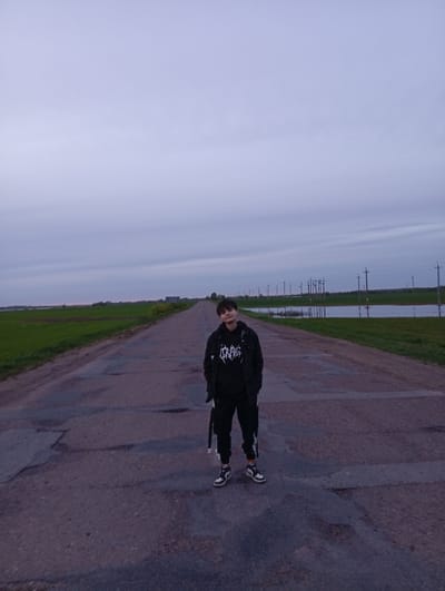 The photograph depicts a person standing on a paved road. The individual has short dark hair, and is wearing a black hooded sweatshirt with a graphic print on the front, black pants, a black jacket, and two-toned shoes. They are facing the camera with their hands in their pockets. The road appears to be cracked and uneven. On either side of the road, there is grass, and what appears to be standing water. In the background, telephone poles stretch into the distance beneath an overcast sky. The location is Azyakul', Russia.