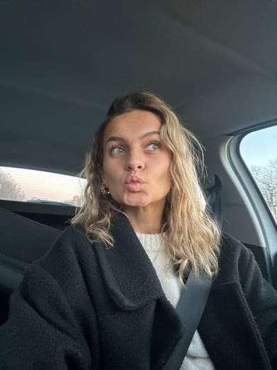 A woman sits in a car wearing a seatbelt and a black coat over a white sweater. She has blonde, curly hair and is making a kissing face toward the camera. The interior of the car is visible, including the ceiling and a window with trees visible outside. The woman is wearing round earrings.