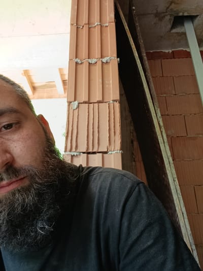 A close-up, half-portrait captures a man at a construction site, likely in Leshnitsa, Bulgaria, during daytime, indicated by the bright natural light. The man's face occupies the left side of the frame, showing his left eye, part of his forehead and cheek, and a significant portion of his thick, dark, and partially graying beard. He wears a dark (likely black) t-shirt and looks directly towards the viewer with a neutral expression.
The background reveals exposed building materials. A vertical structure made of reddish-brown, perforated clay blocks, cemented with visible mortar, forms what appears to be a column or unfinished wall in the center and right of the image. To the left, above the man's head, wooden beams or rafters indicate a ceiling or roof framework. On the right, a dark, long plank or beam leans against the clay blocks, and further right, a lighter-colored, possibly metallic, vertical support is embedded into a concrete ceiling or lintel. Some green foliage is faintly visible in the upper left background, suggesting an opening to the outside. No discernible text is present in the image.