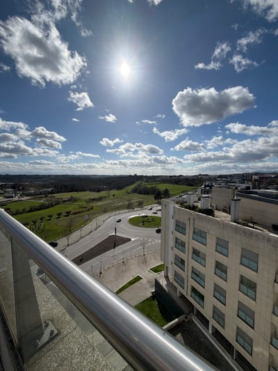 An elevated, outward-looking perspective captures an urban and semi-rural landscape under a bright, partly cloudy sky in Viseu, Portugal. The prominent sun, centered in the upper frame, radiates intensely, accompanied by scattered white cumulus clouds against a clear blue background.
In the immediate foreground, a silver-colored metal railing, indicative of a balcony or terrace, spans horizontally across the lower third of the image, resting on a speckled granite-like surface.
The midground features a significant road network. A large, multi-lane asphalt roundabout, with a central landscaped island, is visible, accommodating multiple vehicles (primarily dark-colored cars) in motion. Connecting roads lead off the roundabout. Adjacent to the roundabout, on the right side of the frame, stands a multi-story, light-colored building with a grid of rectangular windows, characteristic of modern institutional architecture, likely part of the Hospital São Teotónio complex. Its rooftop displays various ventilation units and structural elements.
Beyond the road infrastructure, the midground transitions into an expansive area of vibrant green fields interspersed with numerous trees and darker forested sections. This green space appears well-maintained, suggesting parkland or agricultural use.
The background reveals a distant horizon with further urban structures integrated with additional green areas and what appear to be low-lying hills or mountains. The scene depicts a clear, sunny day, with no discernible human figures interacting directly in the visible portions, save for the occupants of the moving vehicles.