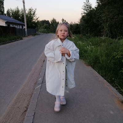 A young blonde-haired female child, with light-colored eyes, is depicted walking on a paved surface in Kryukovo, Russia. She faces directly towards the camera, holding her hands clasped together at chest level. She is dressed in an oversized, light-colored (appearing off-white or light grey) long-sleeved jacket or coat with dark buttons, white tights, and light pink strapped shoes. The paved surface extends to a dirt shoulder and an asphalt road on the left, separated by a concrete curb. To the right of the child, a strip of green grass and foliage contains sparse white flowers. In the left background, a single-story building with a dark roof and light-colored walls is partially visible behind a dark corrugated fence, adjacent to a utility pole. The right background features a dense line of various trees and undergrowth. The sky above is a pale, muted tone, indicating a twilight setting.