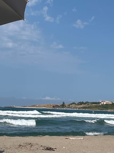 The media file depicts a beach scene with a clear blue sky and scattered clouds. The ocean is on the right side of the image, with prominent white waves breaking. The beach itself is sandy and appears to be somewhat rocky, with debris scattered on the sand. In the background, there is a line of trees and buildings that suggest a coastal town or resort. The image is oriented vertically, with the sky occupying the left half and the ocean and beach on the right. A portion of a dark umbrella is visible in the bottom left corner of the frame. The time of day appears to be daytime, given the bright sunlight and clear sky. There are no people clearly visible in the frame, though there might be a distant figure on the water. The overall impression is of a sunny day at the beach, possibly in a developed coastal area.