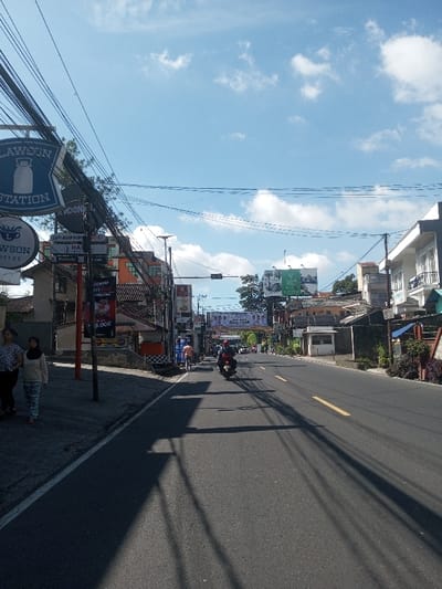 This image captures a wide-angle street scene in the city of Ban Phonhxavanh, Laos, under a bright blue sky with scattered white cumulus clouds. The view is down an asphalt road featuring a yellow dashed center line, with long shadows cast across it by utility poles and overhead wires.

On the left side of the street, a paved sidewalk accommodates two women walking away from the camera. The foreground woman wears a patterned top and dark pants, while the second wears a light-colored long-sleeved top, patterned pants, and a dark head covering. Above and to their left, an extensive network of black utility cables spans the scene. Prominent blue and white signs are visible, one for "LAWSON STATION" featuring a milk bottle graphic, and another below it for "LAWSON COFFEE." Further along the left side, numerous commercial buildings display various banners and signs, with partially legible text including "SAPI ASAP REMPAH NO. 1," "NUSJOLA," "MIE AYAM JAM," and "BAKSO." A large banner stretched across the street, farther in the distance, reads "Selamat Datang... IDUL FITRI."

On the road itself, a motorcycle with two riders, one wearing a red helmet, travels away from the viewer. Other indistinct vehicles are visible further down the thoroughfare. The right side of the street is lined with multi-story buildings of varying architectural styles, some featuring balconies and gray exteriors, interspersed with green vegetation. A large green billboard is situated above these buildings on the right side, further down the road. The overall environment presents a bustling urban commercial corridor during daytime.