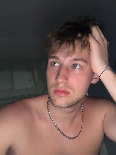 A young man with light brown hair is shown from the chest up, looking to his right. He has blue eyes and is wearing a silver chain necklace. His right hand is raised to his head, and his left arm is resting on his chest. The background is dark and appears to be indoors. The image does not contain any information about the location in the city of Vanino, Russia.