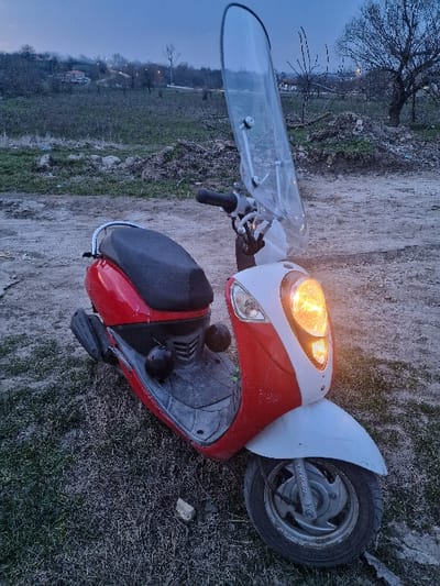 A two-tone red and white motor scooter is stationary on an unpaved, sparsely vegetated surface in Sini Vir, Bulgaria. The scooter's front headlight is illuminated, emitting an amber light. It is equipped with a tall, clear plastic windshield and a black textured seat. Two dark, spherical objects rest on the scooter's floorboard. The immediate ground consists of compacted dirt and dry, patchy grass. The background reveals a gently ascending terrain with deciduous trees, some appearing bare, under a dim, blue-grey sky consistent with twilight conditions. Distant structures, likely residential, are visible on the distant hillside.