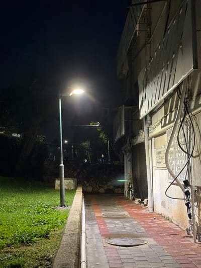 This image depicts an outdoor urban pathway at night in Migdal HaEmeq, Israel. A tall, greenish-blue metallic lamppost with a single luminaire stands prominently on the left, emitting bright light that illuminates the immediate foreground. To the left of the lamppost is a manicured grassy area. A concrete curb separates this grass from a brick-paved pathway on the right. The pathway is constructed from interlocking rectangular bricks, primarily red and gray, and appears damp. Two circular metal manhole covers are embedded in the pathway.
To the right of the pathway, a light-colored building wall rises vertically, adorned with numerous dark cables, white conduits, and electrical boxes. A window with a white metal security grate is visible on this wall. Above the window, a large, slatted or louvred architectural element extends horizontally.
In the mid-ground, beyond the pathway and behind the lamppost, is a low stone or rock retaining wall topped with dense dark vegetation. Further in the distance, approximately two blurry figures are discernible on an unpaved area or path, and another distant light source is visible. The sky overhead is dark. No specific actions or interactions are clearly discernible among the distant figures.