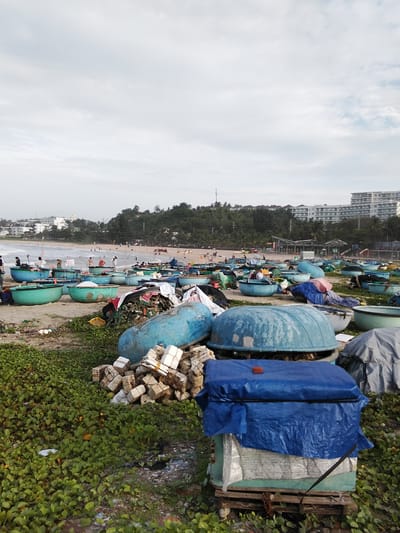 The image depicts a beach scene in Phú Thủy, Vietnam, characterized by numerous round, blue fishing boats clustered on the sand. In the foreground, a pile of foam blocks is stacked near a blue tarp-covered structure. The middle ground features more of the distinctive basket boats, some partially filled with debris, and a wider expanse of beach with more boats. People are scattered across the beach, some near the boats and others closer to the water. The background reveals a treeline bordering a hillside, with several multi-story buildings visible in the distance, suggesting a developed coastal area. The sky is overcast, with a light grey hue indicating a cloudy day. The overall environment is a working fishing beach with a mix of traditional boats and modern infrastructure.