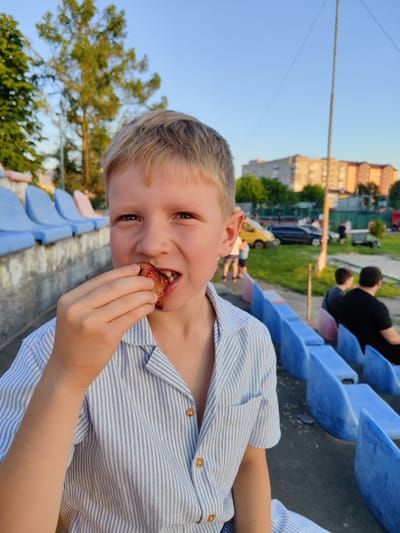 A close-up, eye-level shot shows a Caucasian boy with blond hair and blue eyes, wearing a light blue and white striped button-up shirt. He is sitting in a row of blue and pink stadium seats, holding a piece of food, possibly chicken, to his mouth with his right hand. His mouth is open, and he appears to be chewing. The background is blurred, revealing trees, buildings, cars, and other people seated in the stadium. The lighting suggests it is late afternoon or early evening, with the sun casting a warm glow. The location is identified as the Korosten Dynamo Stadium in Korosten, Ukraine.
