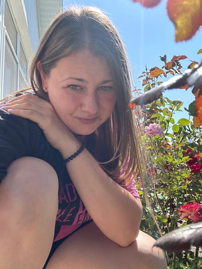 The image shows a close-up of a woman outdoors on a sunny day. She has long brown hair and is looking at the camera with a slight smile. She is wearing a black shirt with pink lettering that reads "BAD GIRL". Her left arm is bent and resting on her shoulder, and her right arm is bent and resting on her leg. In the background, there are rose bushes with pink and red flowers. The scene appears to be in a residential area, with a house visible on the left side of the frame. The woman's skin appears slightly tanned and she has freckles across her nose and cheeks. The lighting suggests it is daytime and the sun is shining brightly.