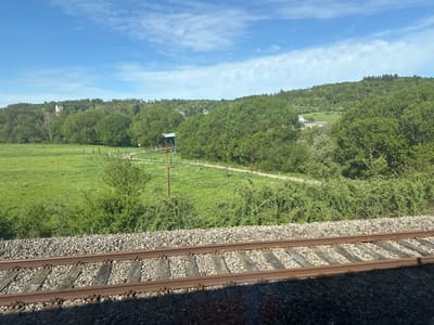 The image displays a serene rural landscape on a bright, sunny day, likely captured from a moving train. In the immediate foreground, the parallel steel rails of a railway track are visible, supported by wooden sleepers set into grey gravel ballast. A dark shadow partially obscures the lower section of the tracks.

Beyond the railway, a wide, lush green field dominates the midground, dotted with numerous small white flowers. A winding dirt path traverses this field, bordered by sparse wooden posts and an abundance of green bushes and small trees. Further along the path, a distinctive wooden bridge with a blue, pitched roof spans what appears to be a small stream or depression.

The background is characterized by rolling hills, densely covered with a variety of trees, predominantly deciduous, with some darker clusters of coniferous trees visible on the higher slopes. Several buildings are nestled within this verdant landscape. On a distant hill to the left, a notable white building featuring a dark roof and a prominent central tower or spire stands out. Towards the right, another cluster of structures, some with yellowish walls, can be seen amidst the trees. The sky above is a clear, bright blue, adorned with scattered, wispy white clouds.

The scene conveys a sense of peacefulness, with no direct human activity visible. The strong, clear light suggests it is late morning or early afternoon, indicating pleasant weather conditions. The architectural style of the distant buildings and the overall natural setting are consistent with a European countryside, aligning with the location context of Schieren, Luxembourg. No discernible text is visible within the image.